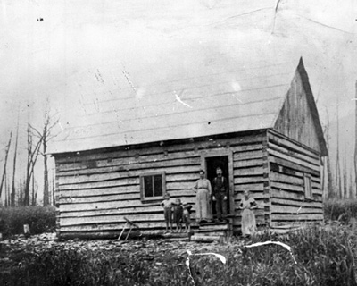 Reproduction d’une photographie de G&eacute;d&eacute;on Bradette et de sa famille sur le seuil de leur maison en bois &eacute;quarri &agrave; queue d’aronde, Cochrane (Ontario), 1909.